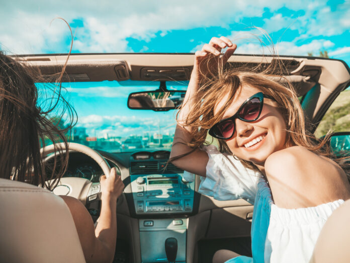 Portrait of two young beautiful and smiling hipster female in convertible car. Sexy carefree women driving cabriolet. Positive models riding and having fun in sunglasses